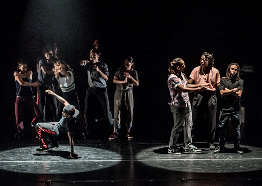 Two pools of light onstage: On the left, one of The Hood Lockers, hand on the ground, feet hovering off the floor, is surrounded by another group of dancers. On the right, the three dancers make a tableau displaying an argument. 
